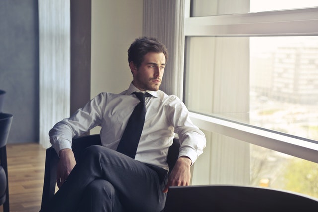 Mladý muž v bielej košeli, tmavých nohaviciach a kravate sedí na stoličke v kancelárii a pozerá von oknom.jpg